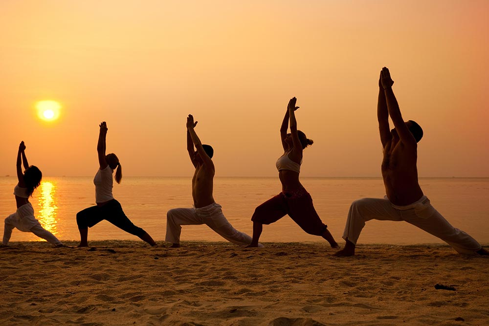 beginningstreatment-what-is-refuge-recovery-article-photo-five-people-practising-yoga-at-the-beach-warrior-i-pose-227779006