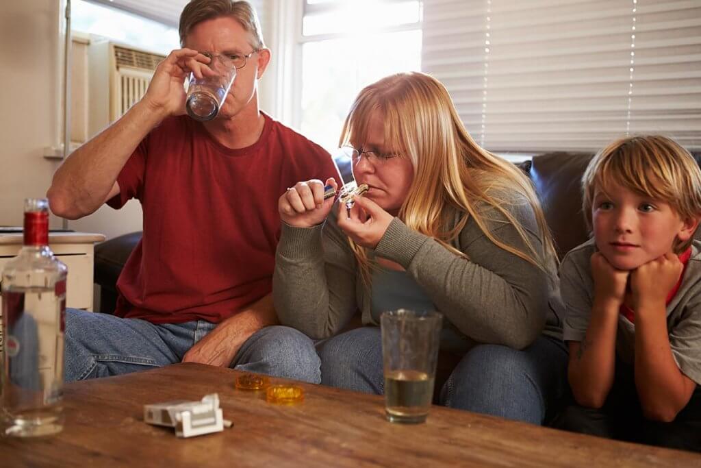 beginningstreatment-child-parent-share-addiction-article-photo-parents-sit-on-sofa-with-children-taking-drugs-and-drinking-193821536